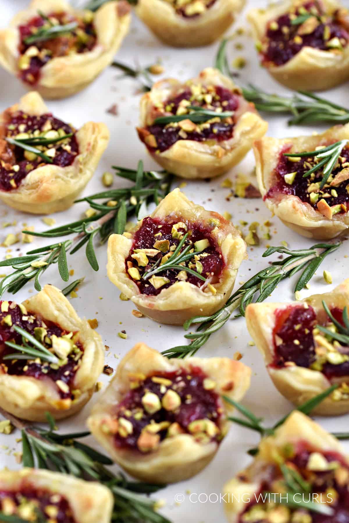 A platter of Puff Pastry Cranberry Brie Bites topped with fresh rosemary and chopped pistachios. 