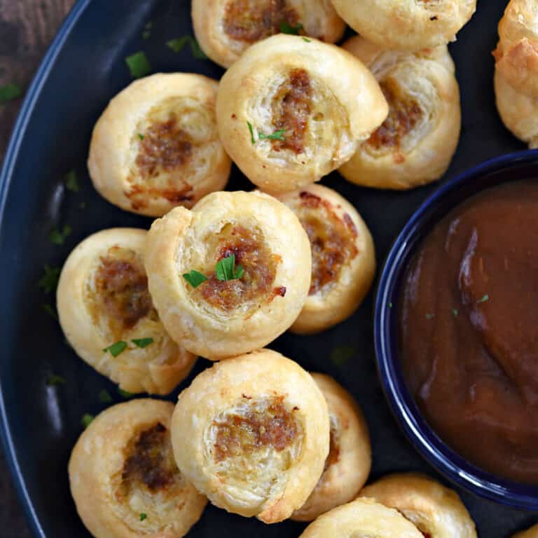 Looking down on a stack of puff pastry sausage bites on a large plate with a bowl of dipping sauce in the center.