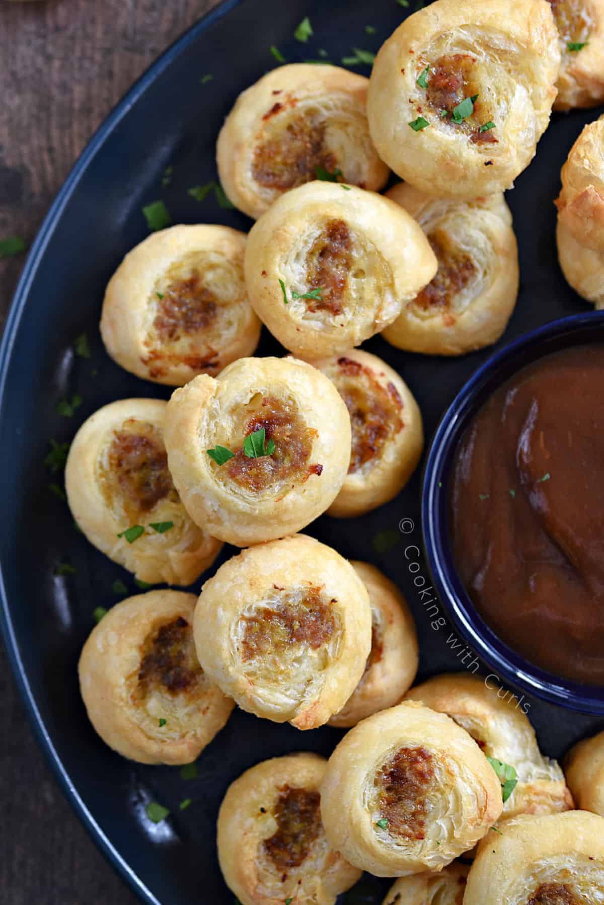 Looking down on a stack of puff pastry sausage bites on a large plate with a bowl of dipping sauce in the center.