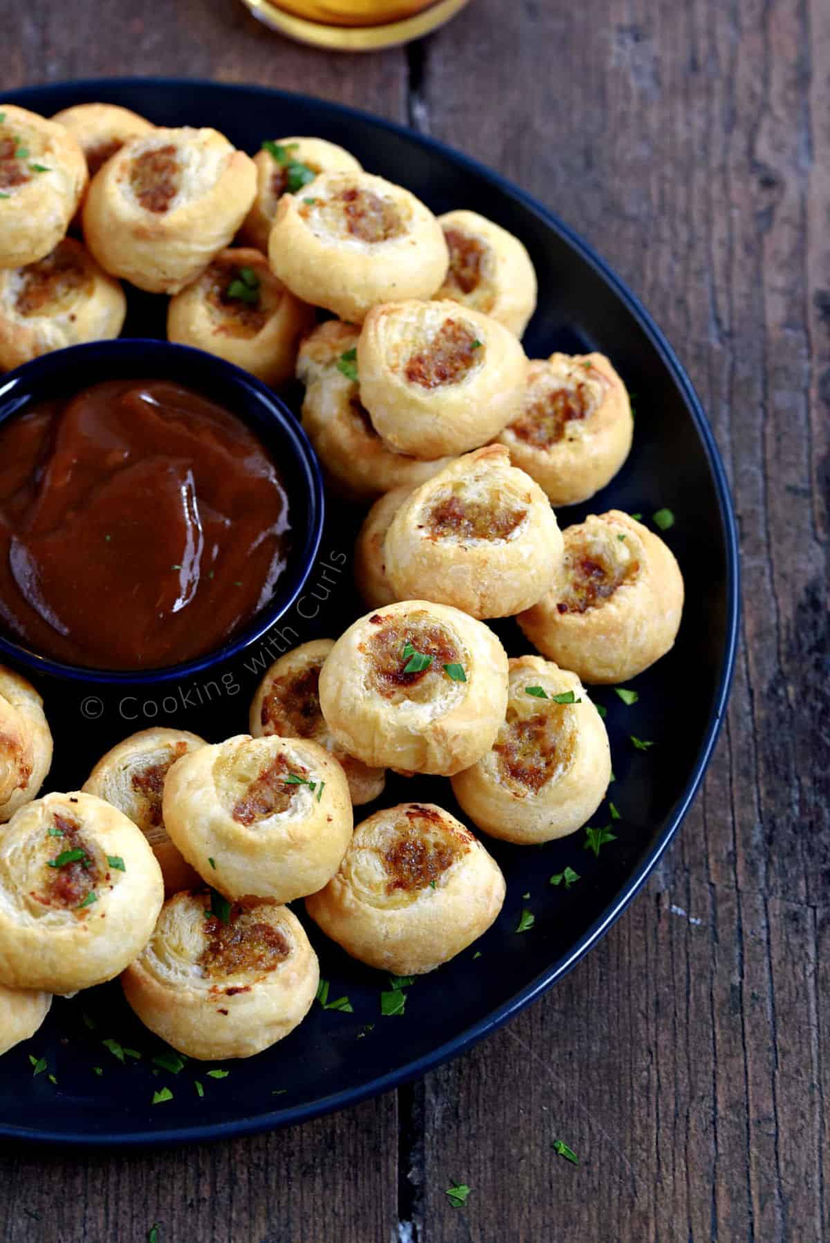 Puff pastry sausage bites stacked on a large plate with a bowl of dipping sauce in the center.