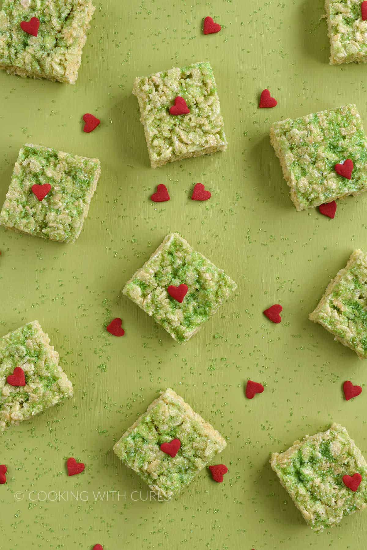 Ten sugar topped rice krispie treats on a green board surrounded by green sugar and red hearts.