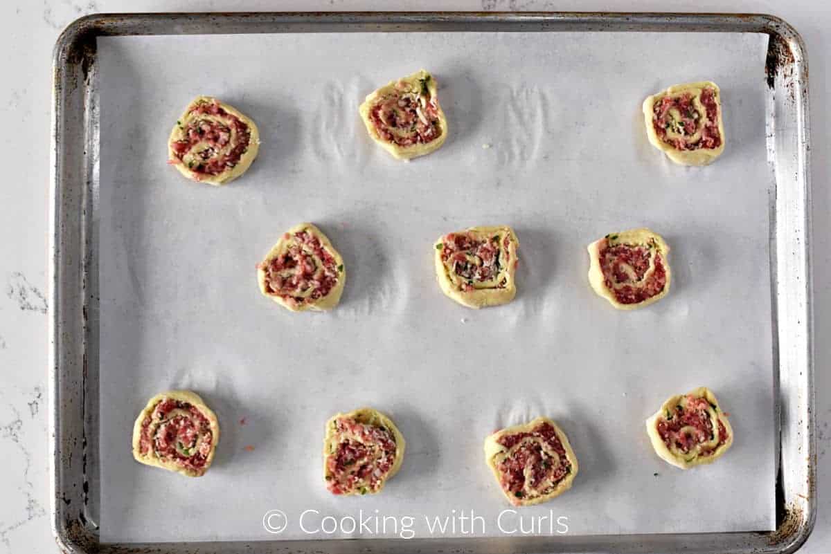 Ten sausage pinwheels slices on a parchment lined baking sheet. 