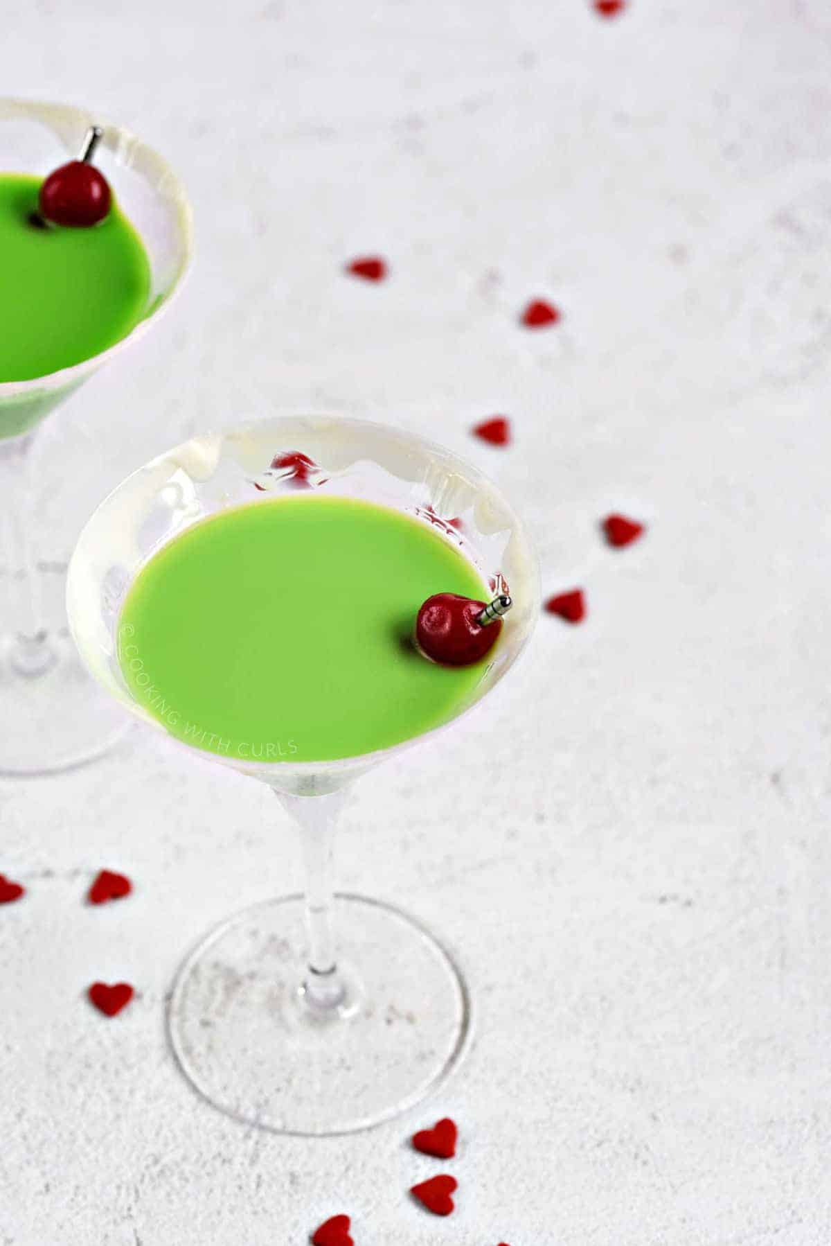 Two martini glasses filled with a green Grinch vodka martini with a cherry garnish surrounded by tiny red heart candies.