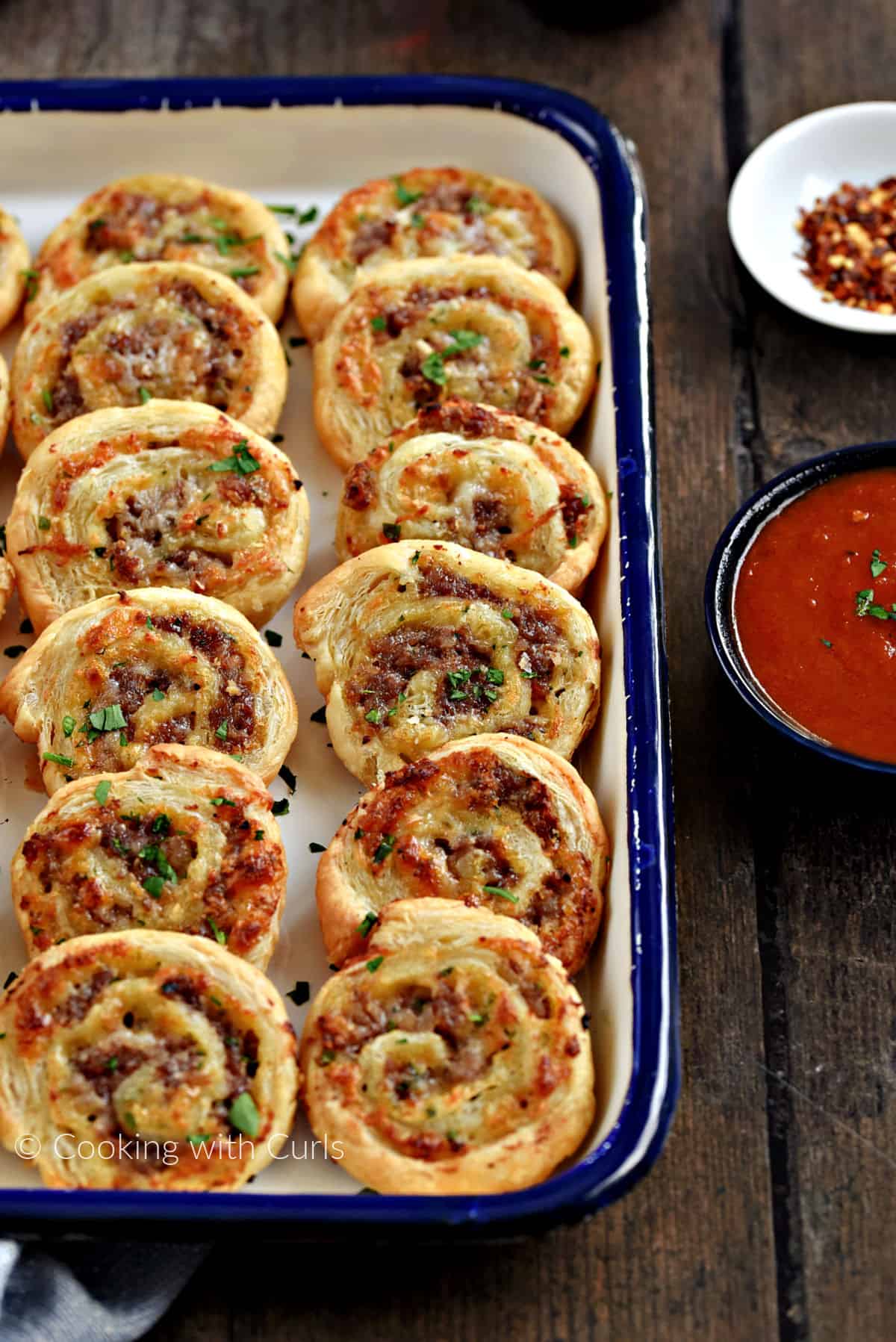 Twelve Sausage Pinwheels on a platter with red pepper flakes and pizza sauce on the side. 