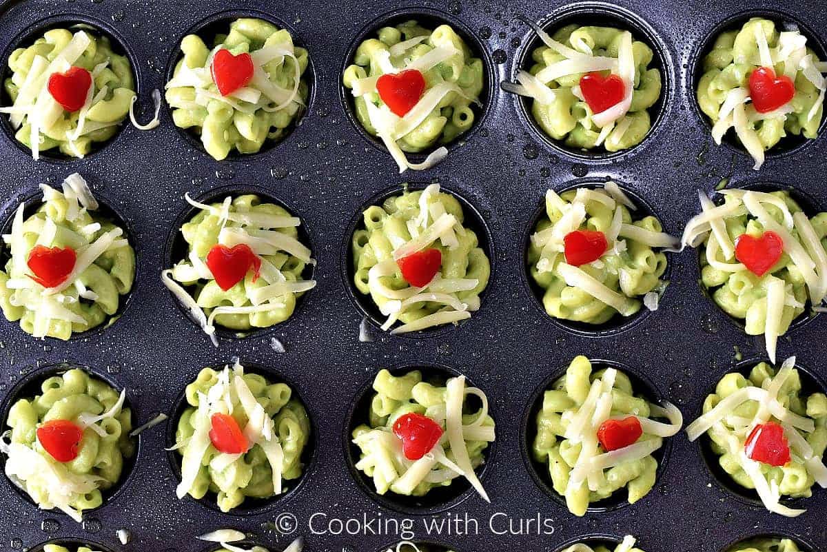 Grinch Mac and Cheese Bites in a mini muffin pan topped with shredded cheese and red pepper hearts.  