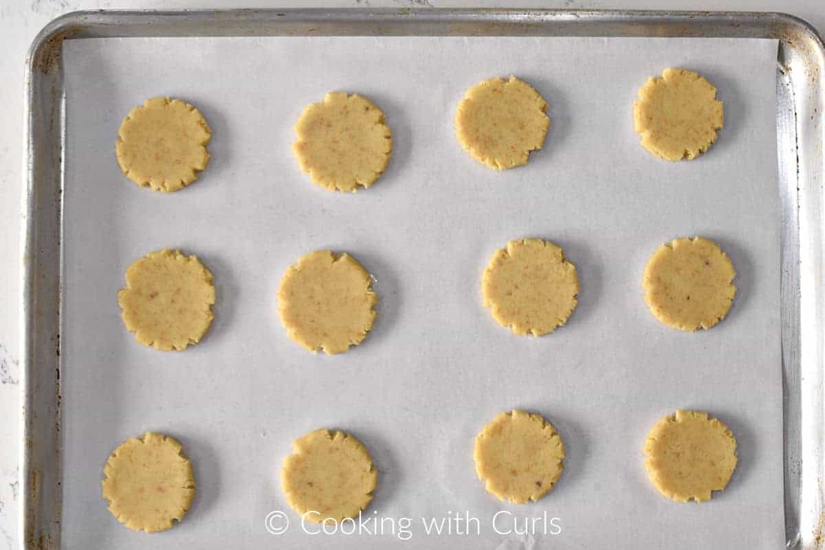 Twelve flattened almond cookies on a parchment lined baking sheet. 