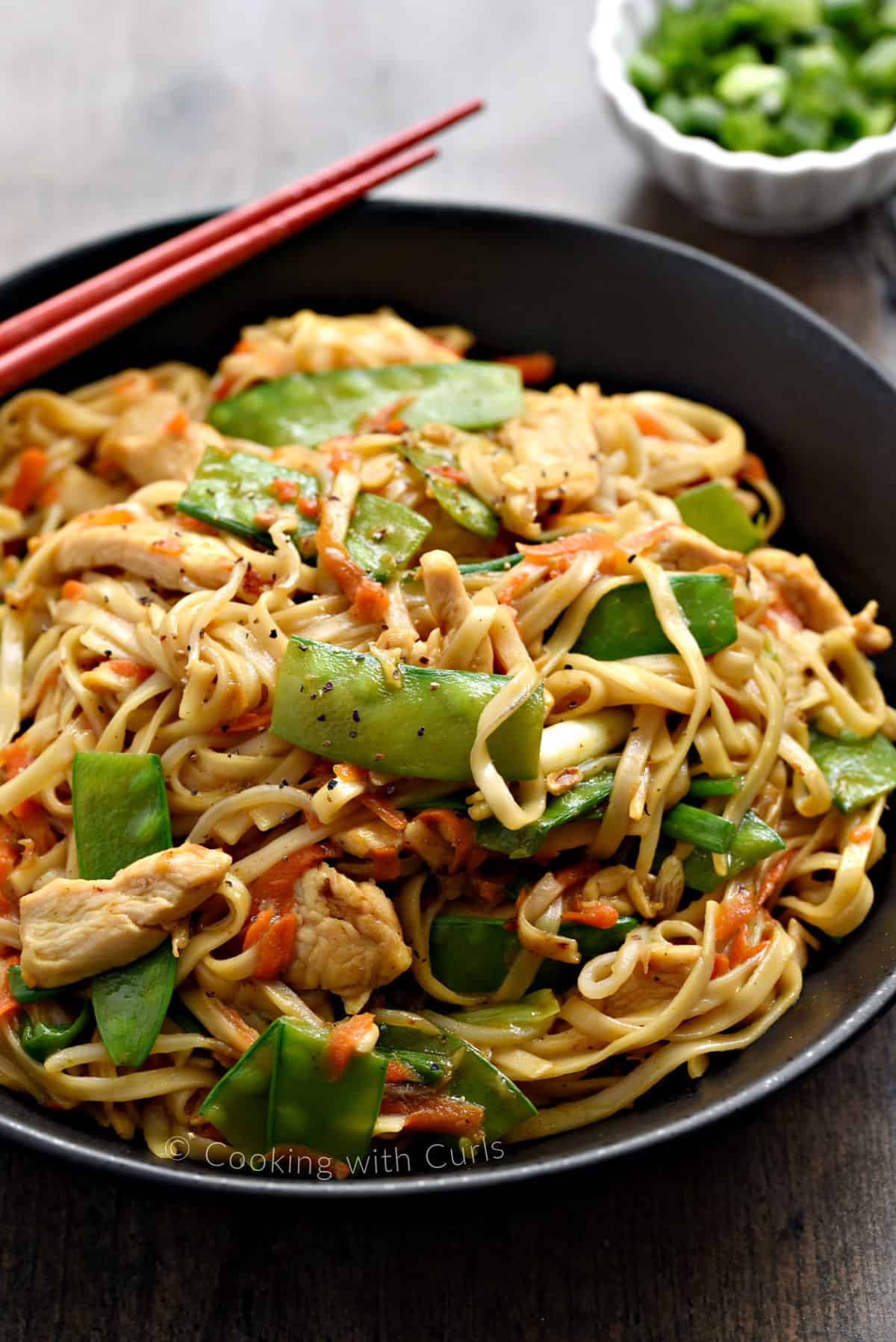 Close-up image of noodles, chicken strips, snow peas, and shredded carrots in a large bowl.