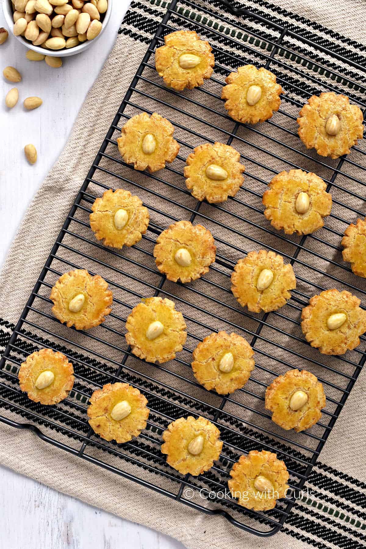 Nineteen Chinese almond cookies on a wire cooling rack over a dish towel with a bowl of raw almonds in the upper left corner.