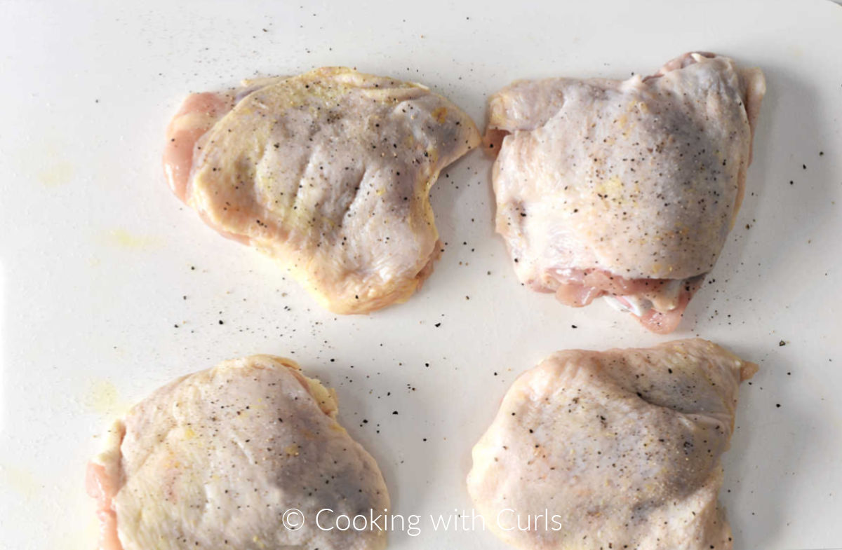 Four raw chicken thighs sprinkled with salt and pepper on a white cutting board.