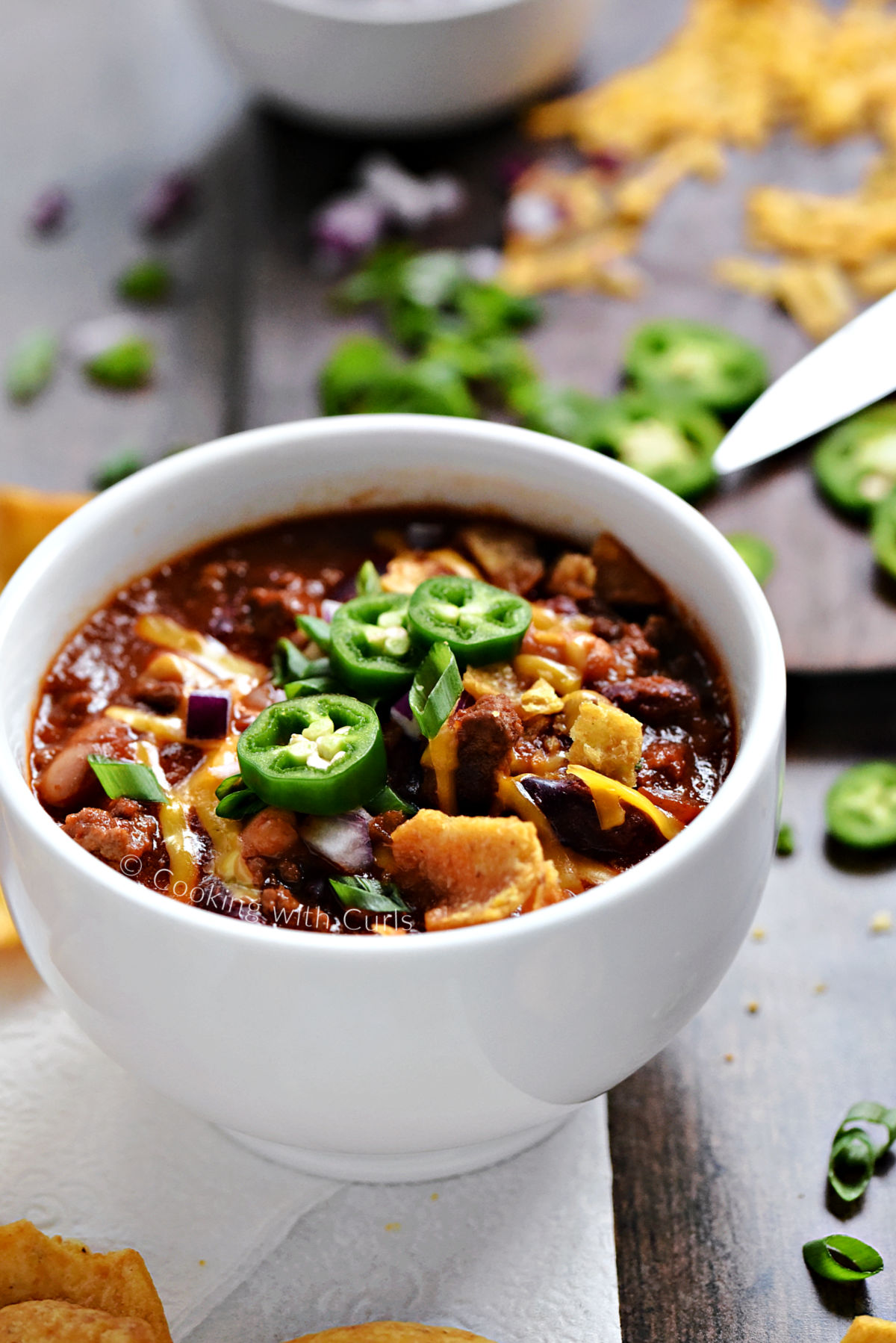 A bowl of Guinness Beer Chili topped with crushed corn chips, melted cheddar cheese, sliced green onions and sliced jalapeños. 
