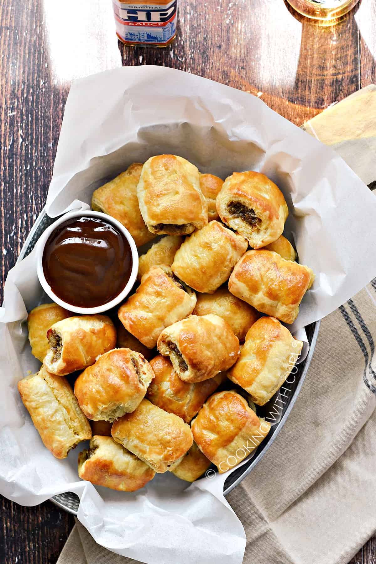 Looking down on a pile of puff pastry sausage rolls in a parchment paper lined basket with dipping sauce in a bowl on the left side.