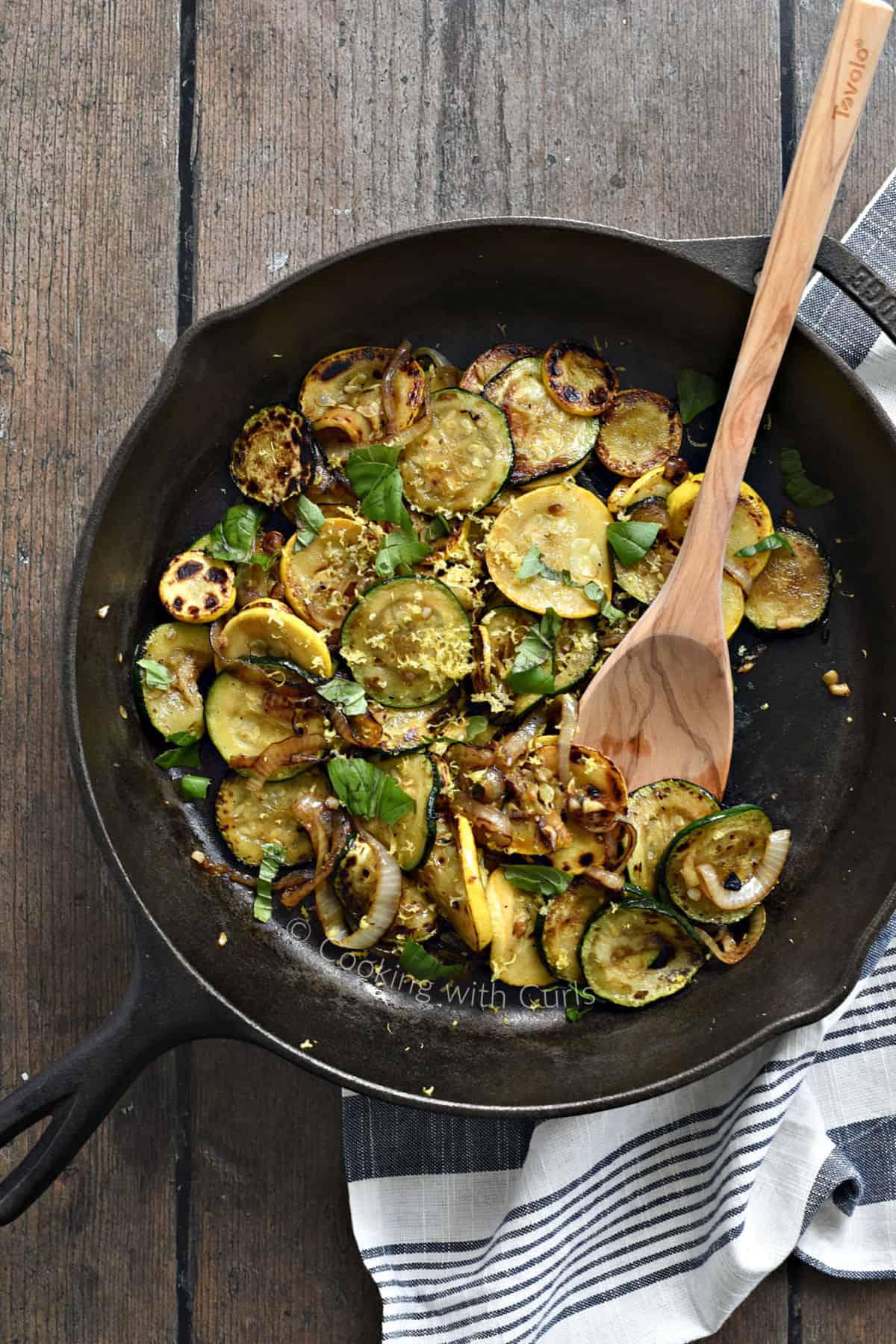 Looking down on sautéed zucchini, yellow squash and onions in a cast iron skillet with a wooden spoon on the right hand side. 