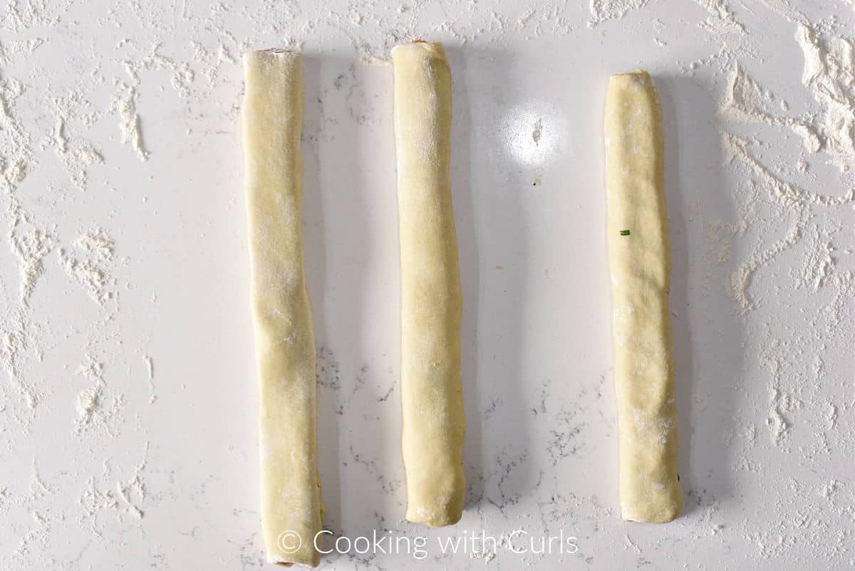Three logs of puff pastry surrounding chicken sausage filling on a marble board. 