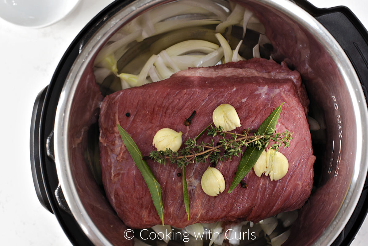 Corned beef, four garlic cloves, peppercorns, cloves, three bay leaves, and sprig of thyme on a bed of onion slices in a pressure cooker. 