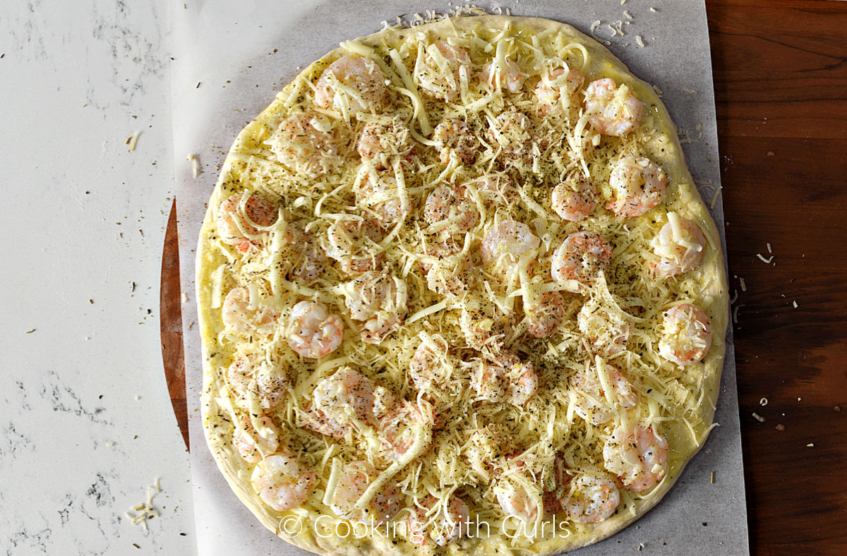 Pizza dough topped with sauce, shrimp, shredded mozzarella and grated parmesan cheese on a sheet of parchment paper. 