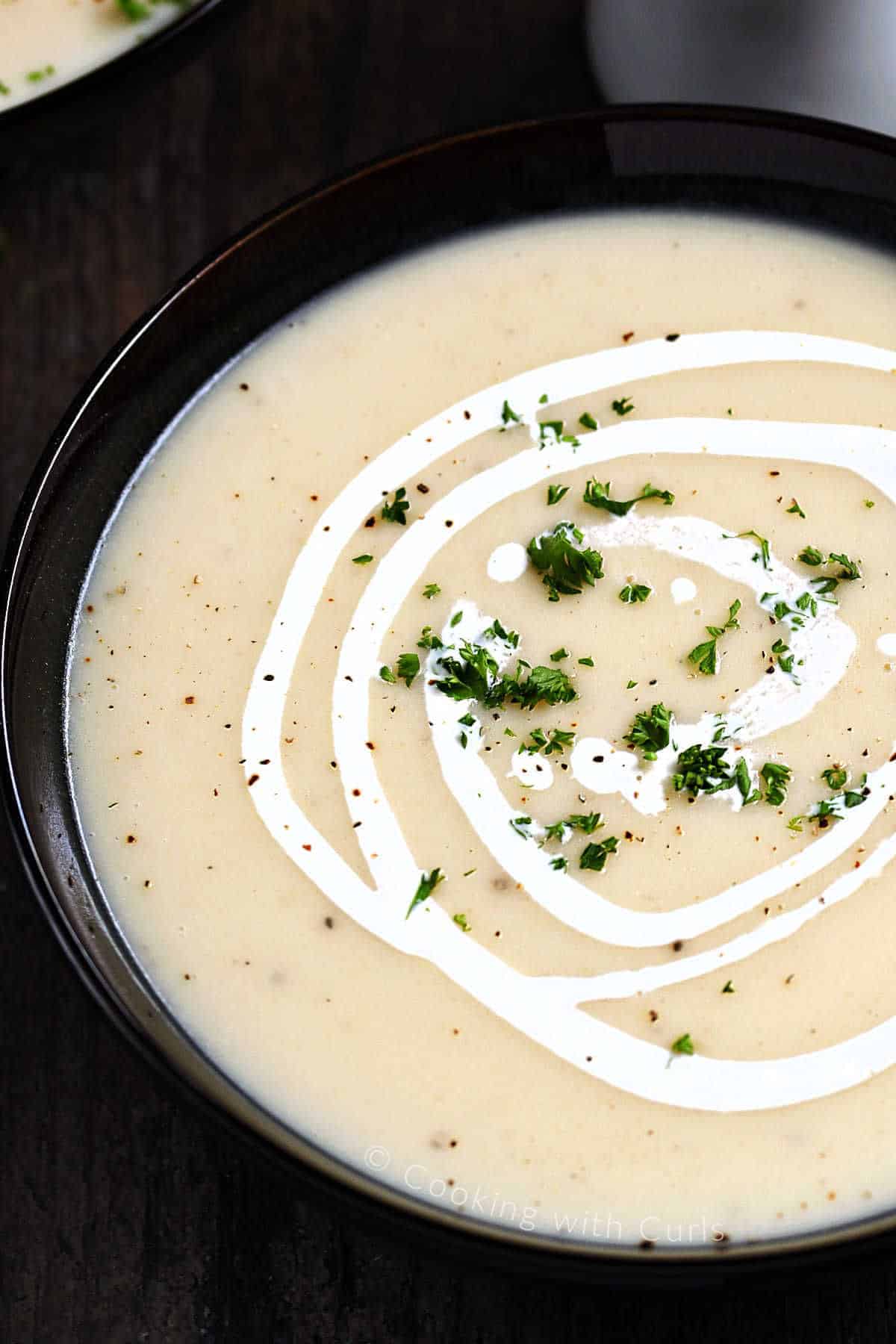 Close-up image of potato leek soup drizzled with heavy cream and sprinkled with minced parsley in a dark bowl. 