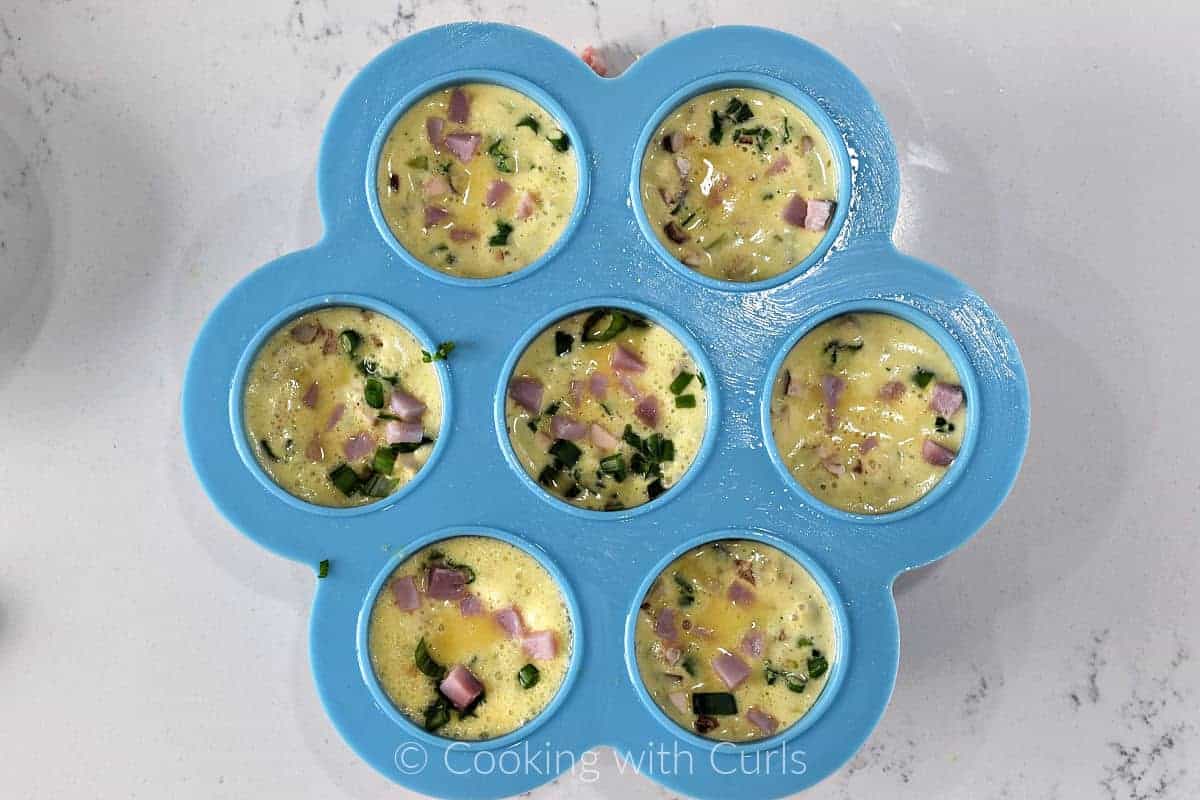 Seven cavities of a silicone mold filled with omelet toppings and egg mixture. 