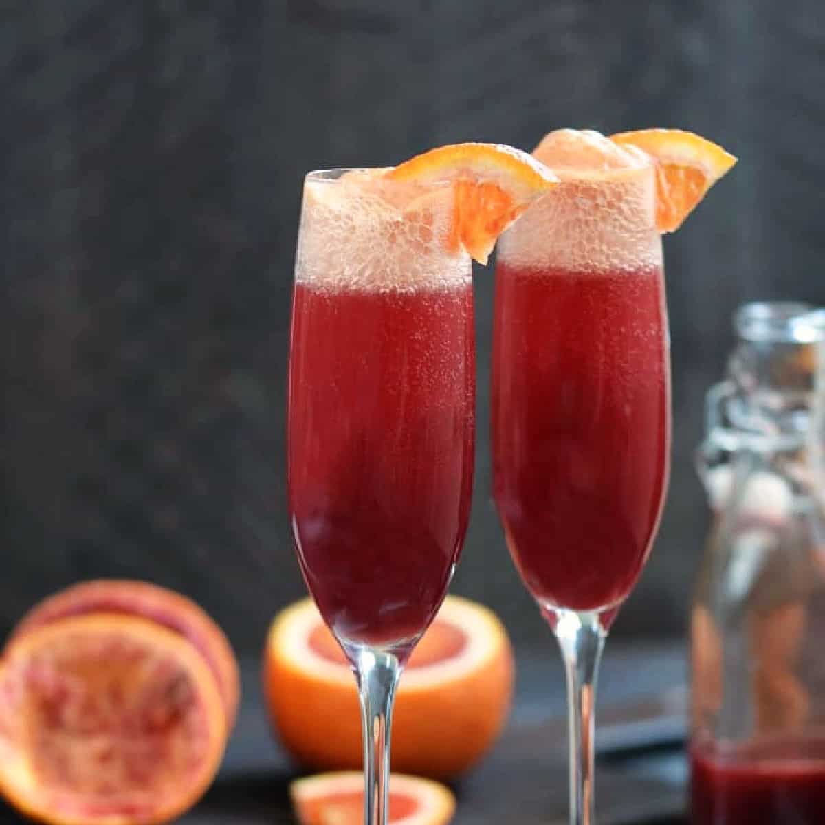 Two champagne flutes filled with blood orange mimosa with an orange garnish.