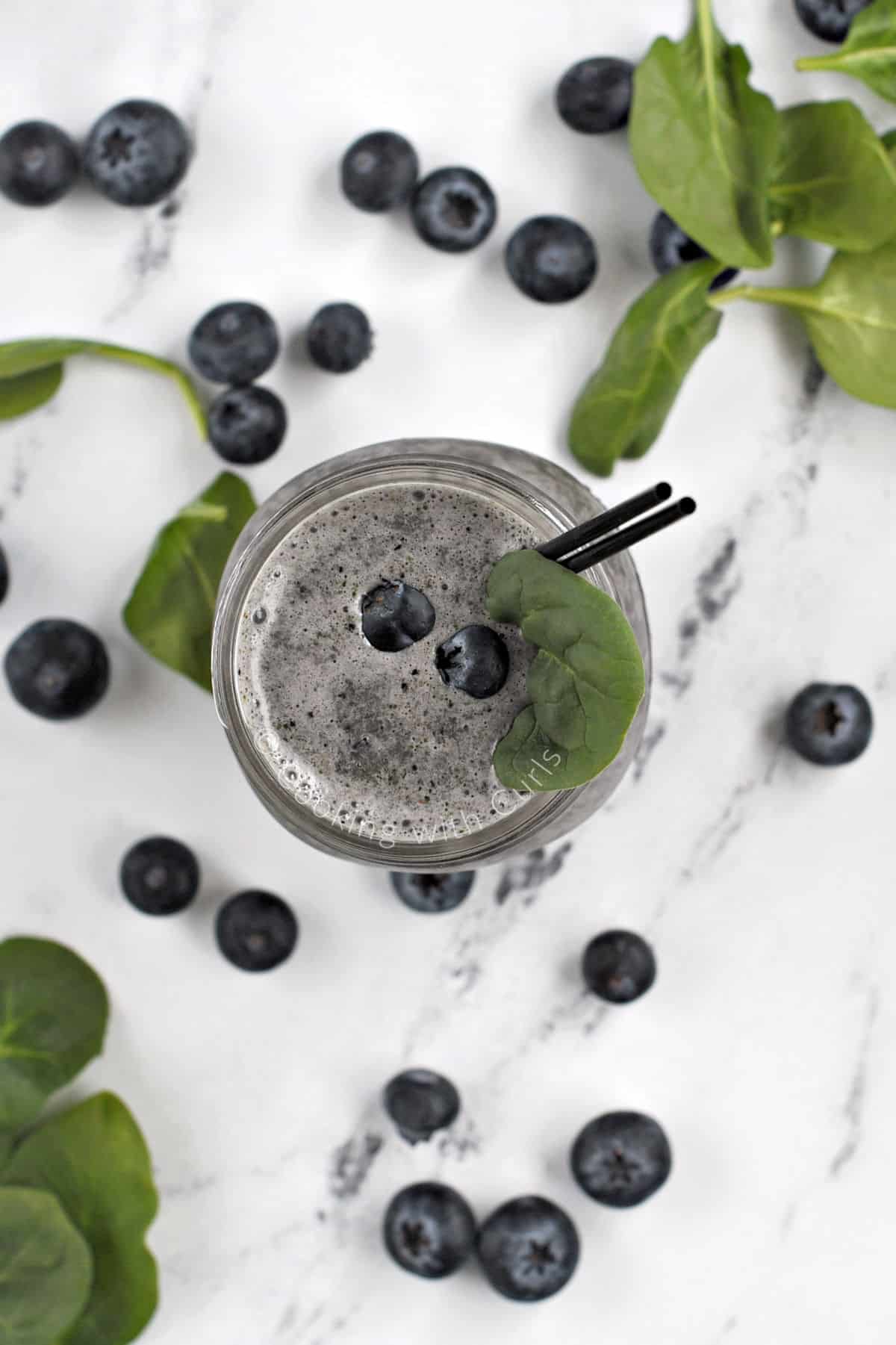 Looking down on a blue smoothie in a glass jar surrounded by fresh blueberries and spinach leaves.