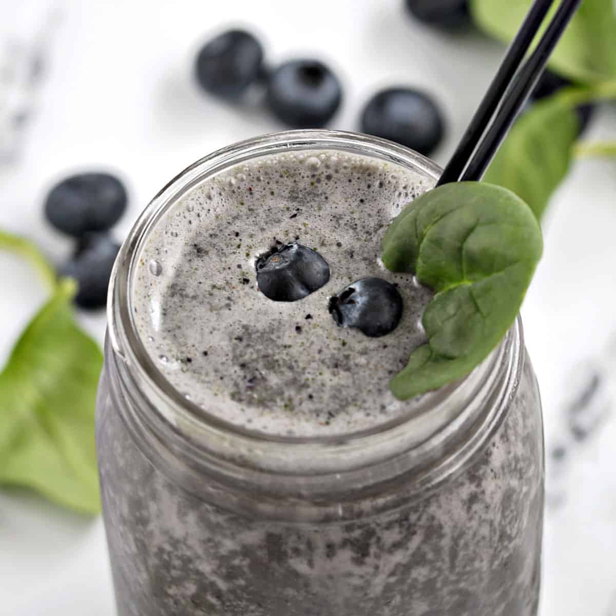 A dark blue smoothie in a mason jar topped with 3 blueberries, a spinach leaf and two skinny black straws.
