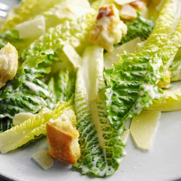 Romaine lettuce leaves topped with Caesar dressing, shaved parmesan and croutons on a dinner plate.