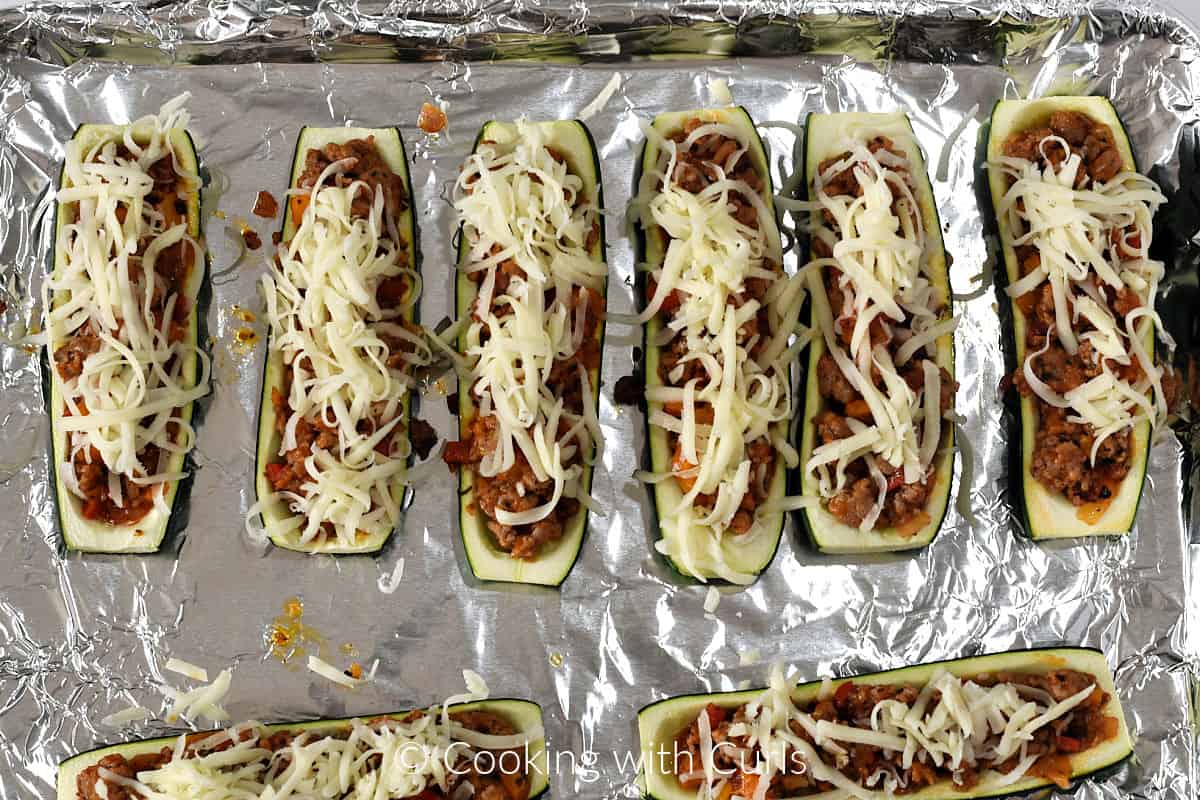 Eight stuffed zucchini boats topped with grated mozzarella cheese on a foil lined baking sheet. 