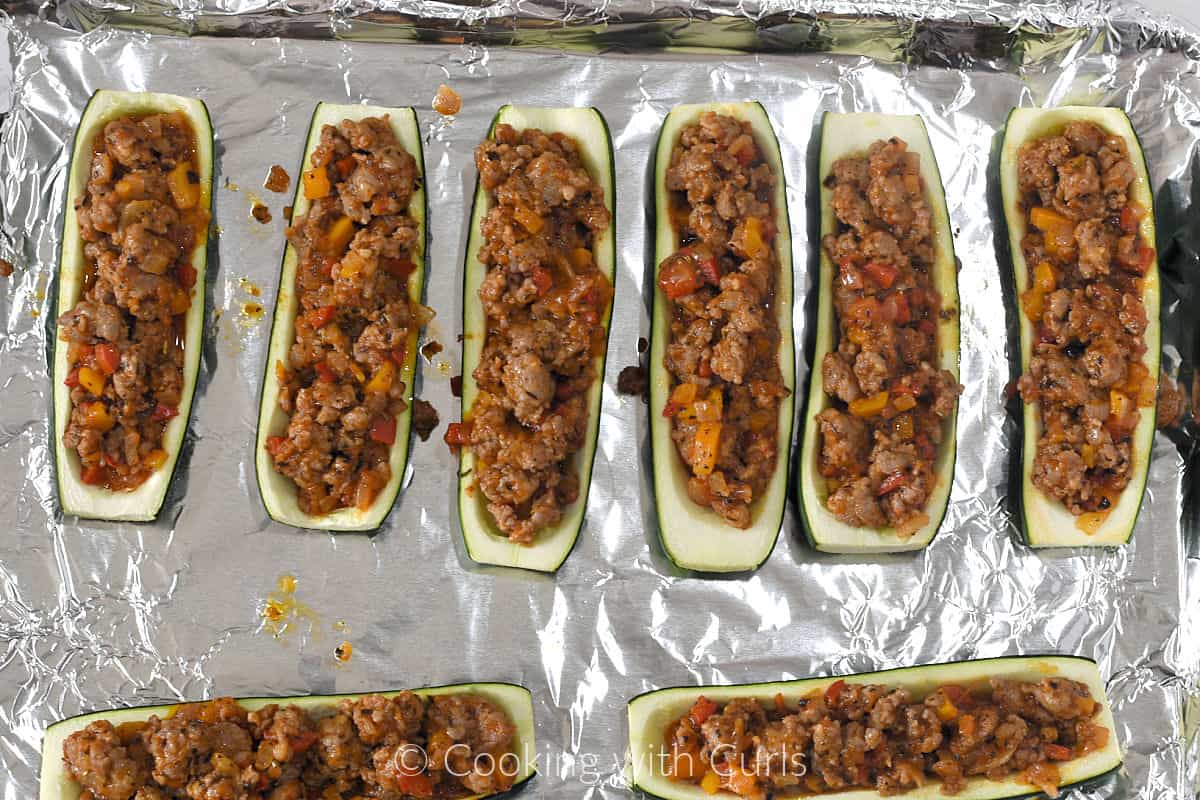 Eight zucchini halves filled with Italian sausage mixture on a foil lined baking sheet. 