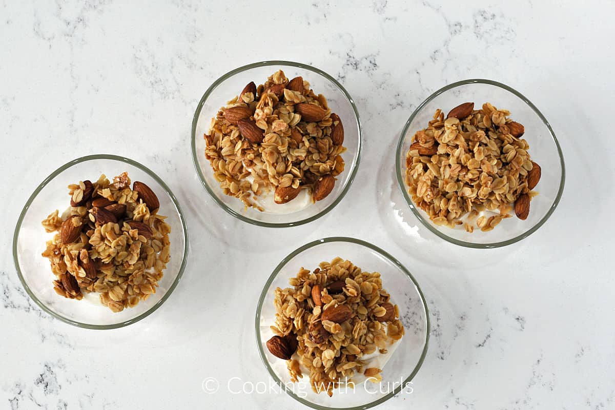 Four small glass bowls with granola on top of yogurt.