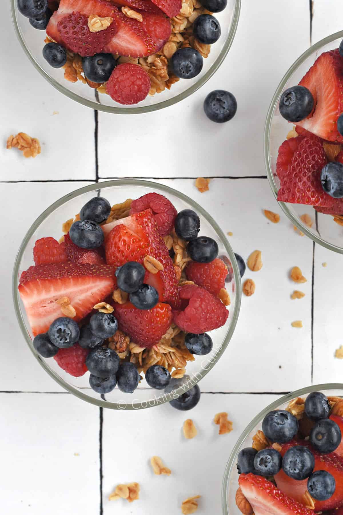 Looking down on four small glass bowls filled with yogurt, granola, blueberries, raspberries and sliced strawberries.