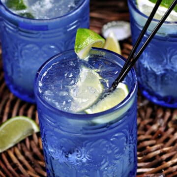 Three blue glasses filled with ice, tequila, lime juice and mineral water with lime wedge garnish.