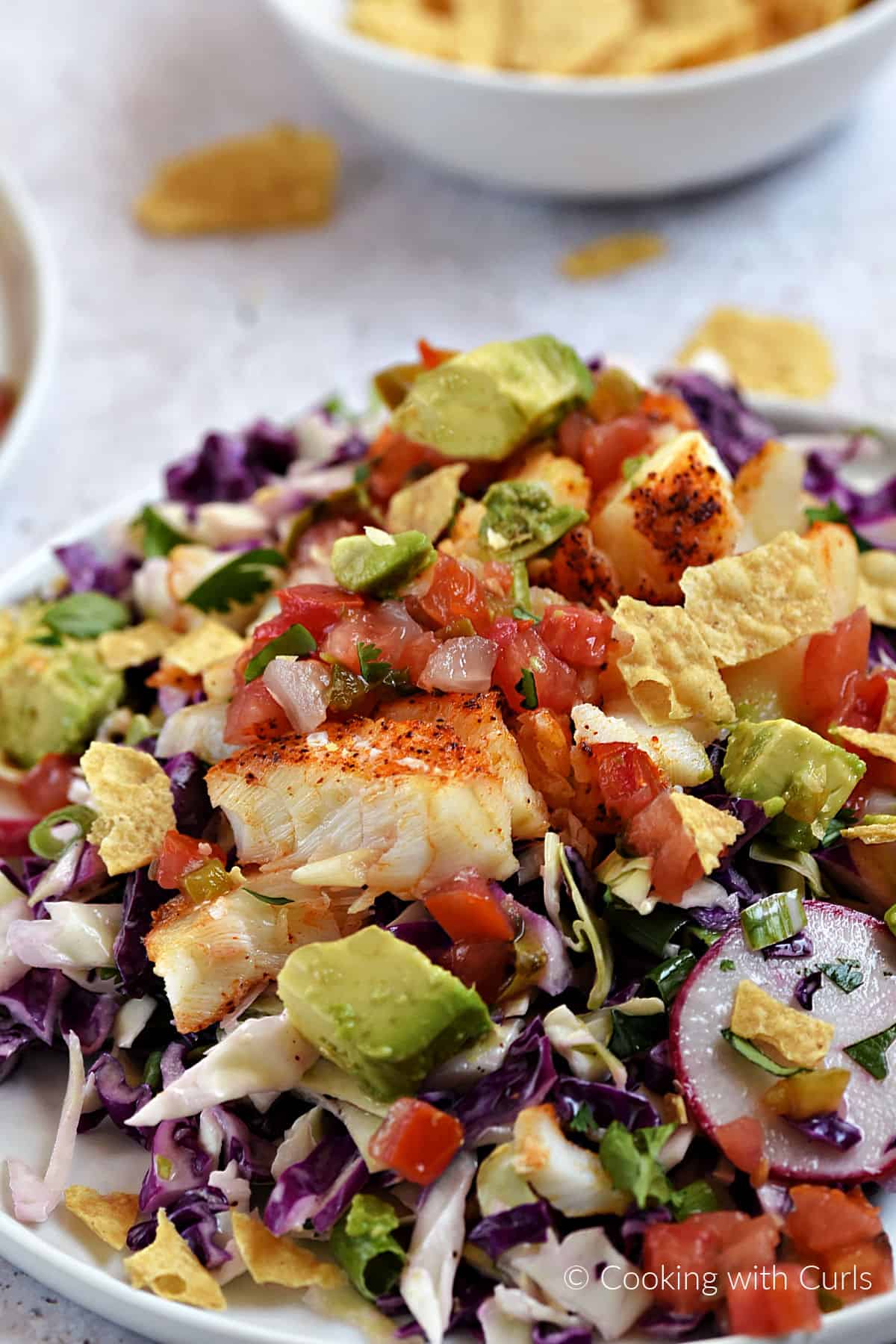 Seasoned cod on top of shredded cabbage with chopped tomatoes and avocado and crushed tortilla chips on a dinner plate.