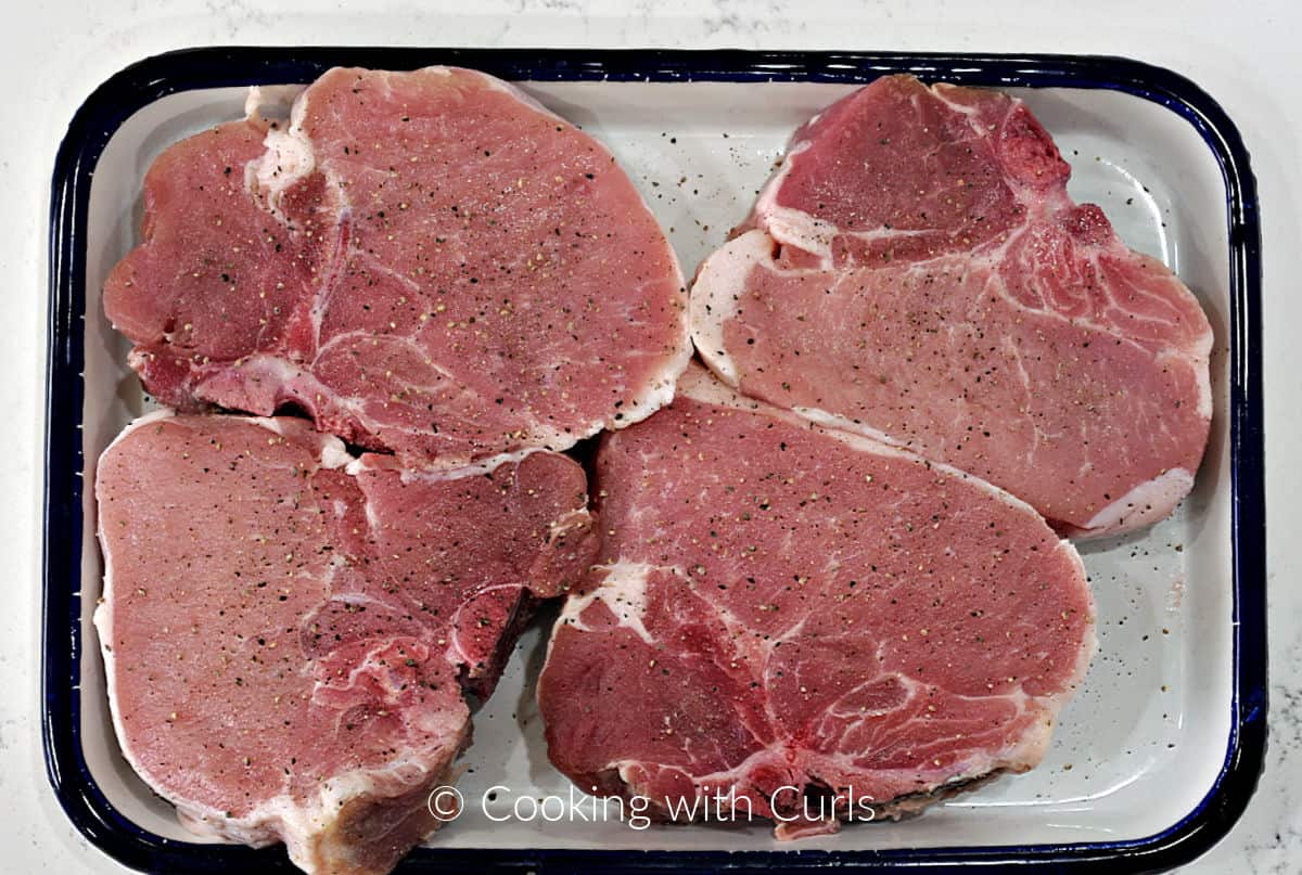 Four thick-cut, bone-in pork chops on a tray seasoned with salt and pepper. 