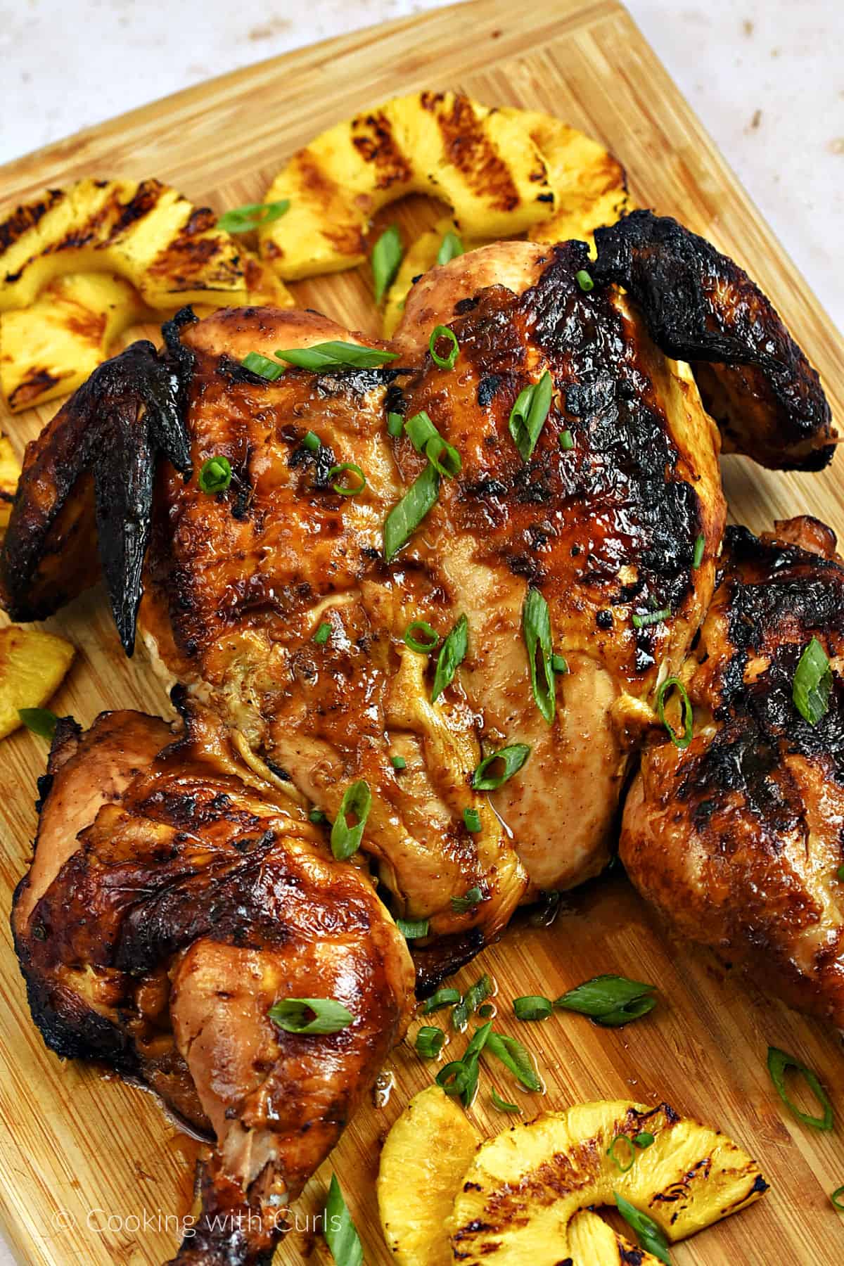 A whole grilled chicken on a wood board surrounded by grilled pineapple slices.