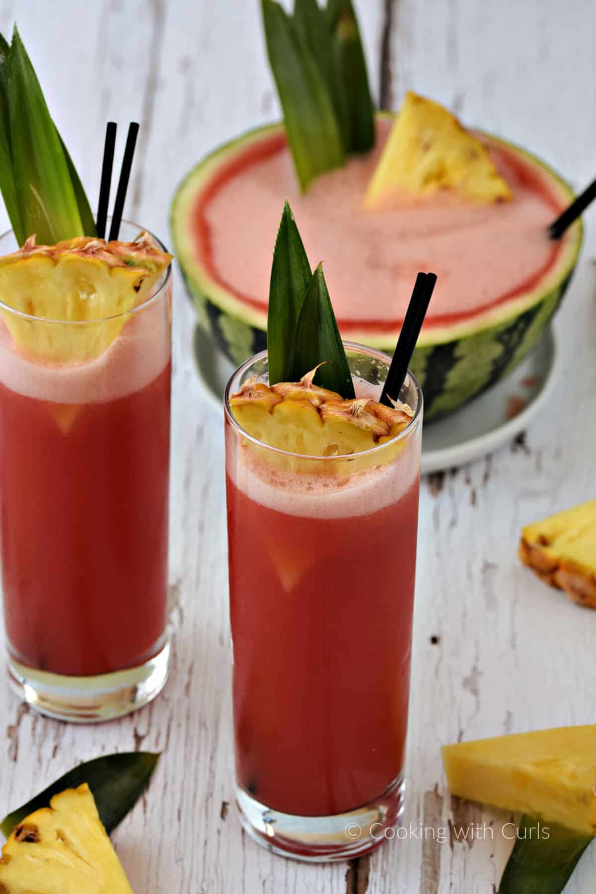 Bright pink punch served in two tall glasses garnished with pineapple leaves and wedges and a filled, hallowed out mini watermelon in the background.