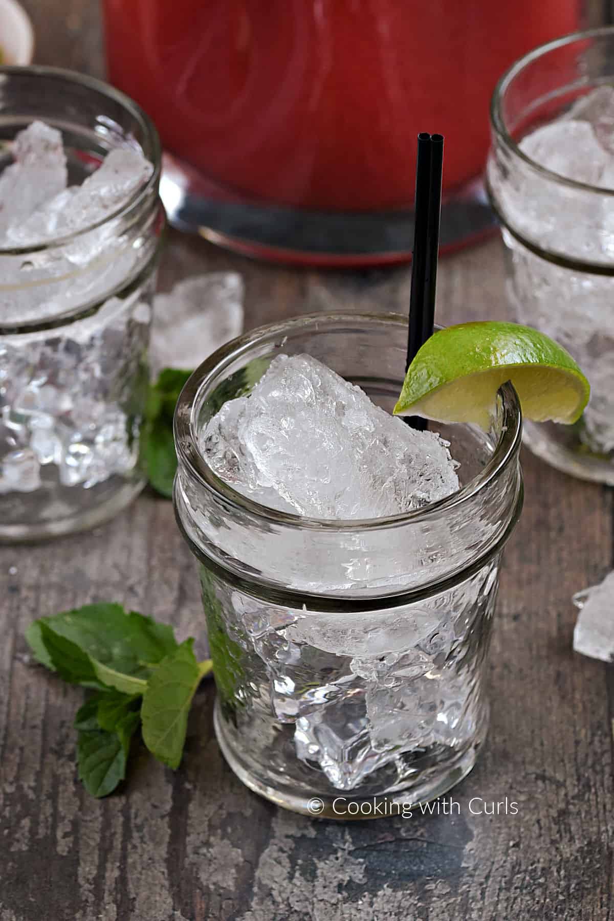 Three cocktail glasses filled with ice chunks with a lime wedge and two black cocktail straws. 
