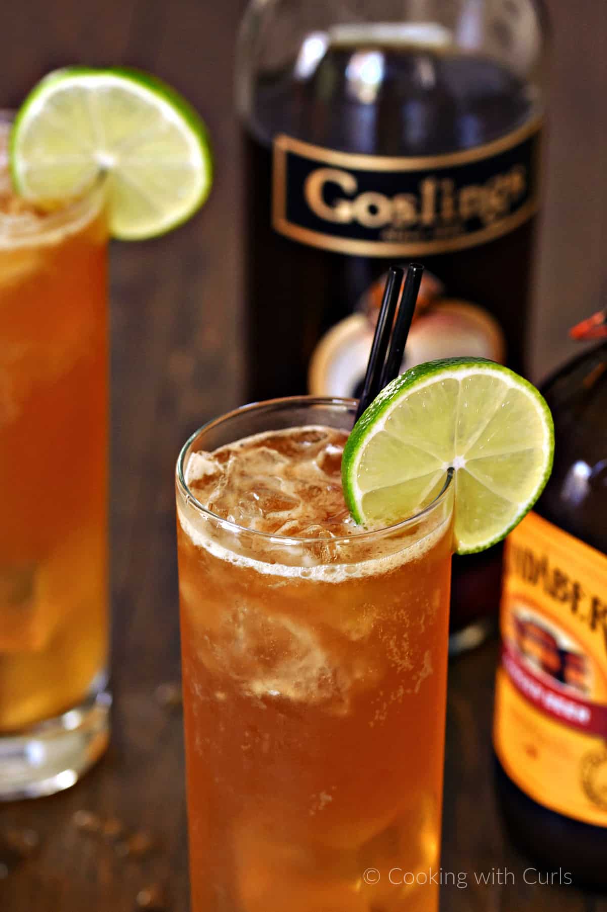 Two tall glasses filled with a bubbly cocktail and a lime wheel with a bottle of goslings dark rum and ginger beer in the background. 