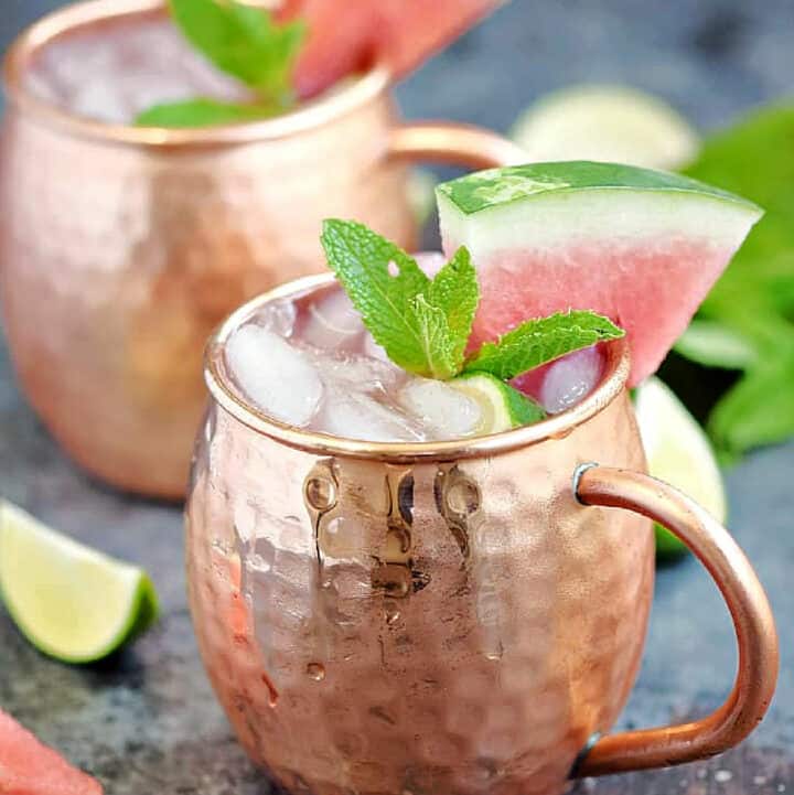 Two copper mugs filled with ice , ginger beer, watermelon juice, and vodka with watermelon wedge and mint leaves garnish.