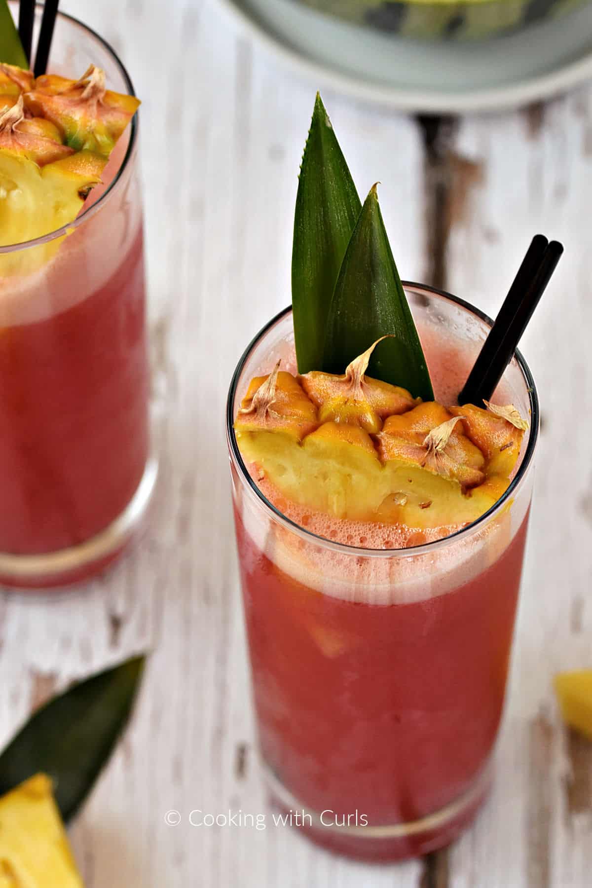 Bright pink punch served in tall glasses garnished with pineapple leaves and wedges.