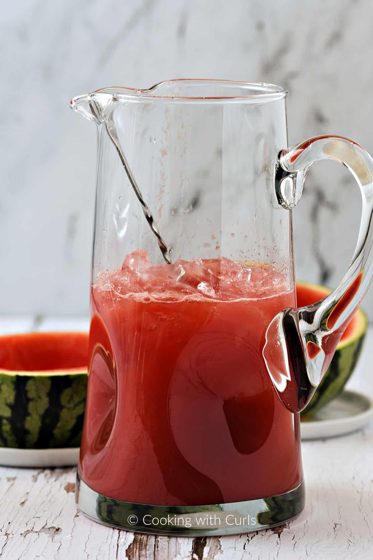 Watermelon juice in a large glass pitcher with ice, rum, orange liqueur, and juices. 