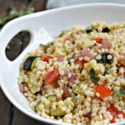 A serving bowl filled with Israeli couscous tossed with roasted red pepper, zucchini, and red onion recipe.
