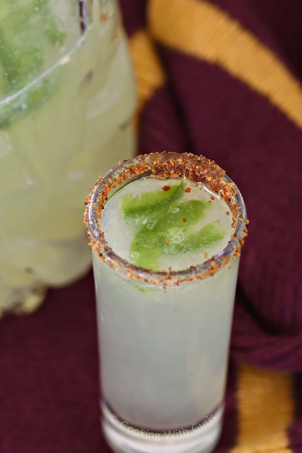 A chili pepper rimmed shot glass filled with spicy jalapeno margarita with a pitcher in the background. 