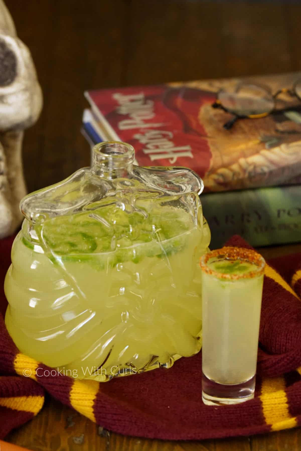 A skeleton shaped decanter filled with spicy jalapeno margarita in front of a stack of Harry Potter books with a filled shot glass in front. 