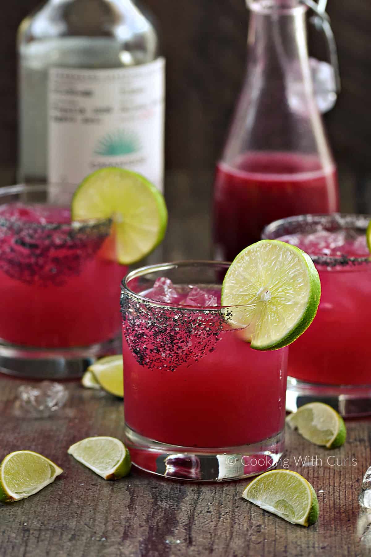 Three prickly pear margaritas in rocks glasses with a bottle of tequila and bottle of syrup in the background.