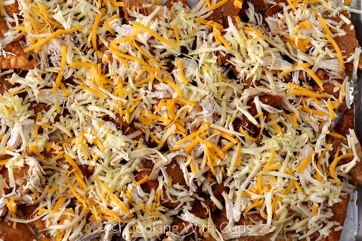 Shredded Jack and cheddar cheese on top of shredded chicken and fried wonton chips on a baking sheet. 