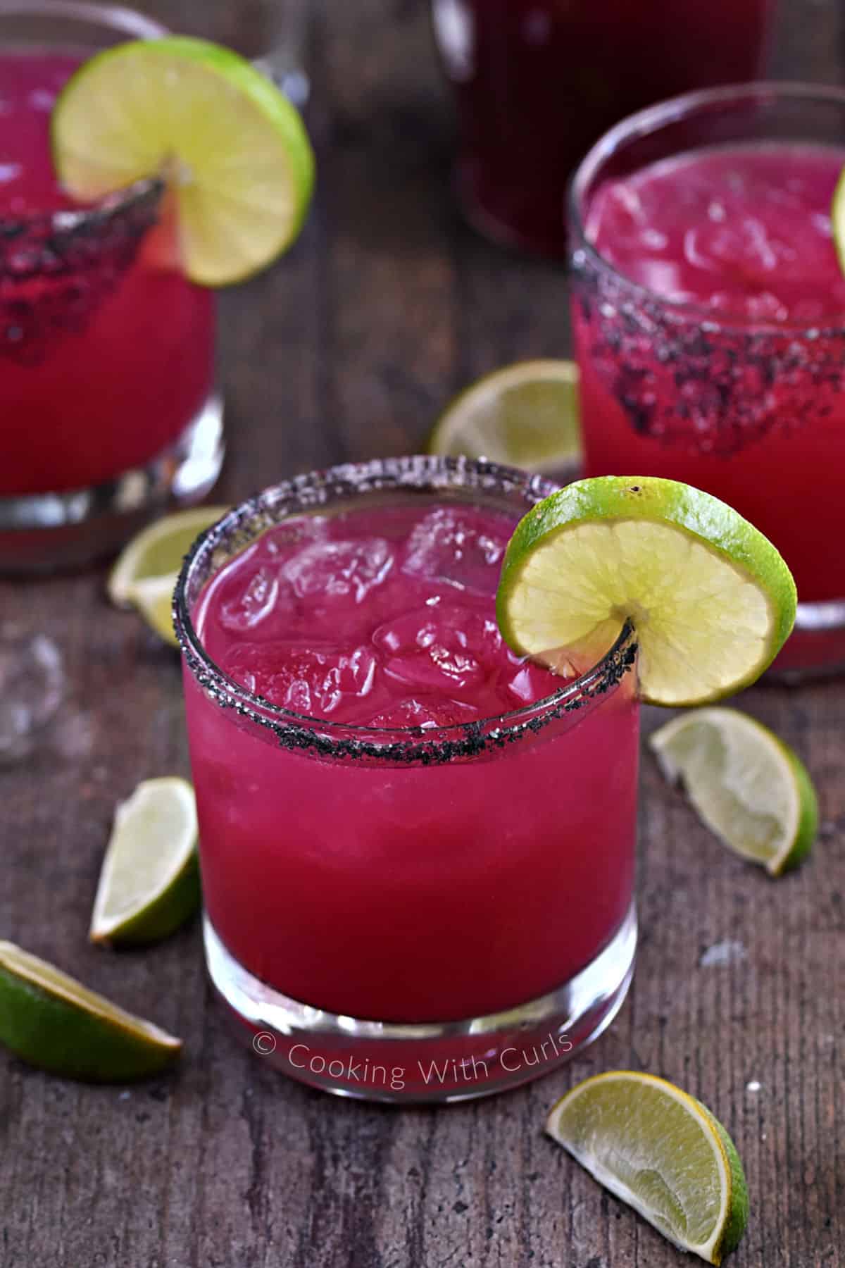 Three rocks glasses filled with bright pink prickly pear margarita with lime wedges and black salted rim. 