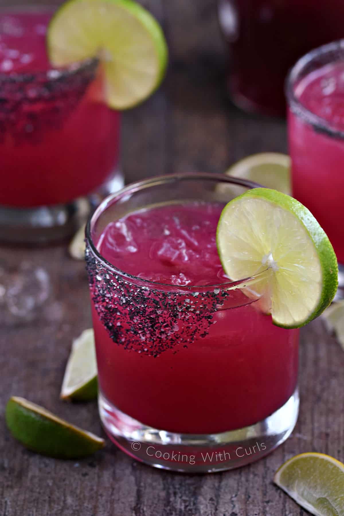 Three short glasses filled with bright pink prickly pear margarita with black salted rim and lime wheels for garnish.