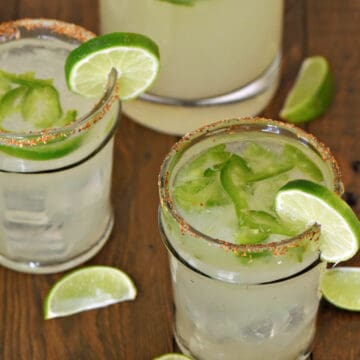 Two ice filled glasses and a pitcher with sliced jalapenos, margarita cocktail, and lime wheels.