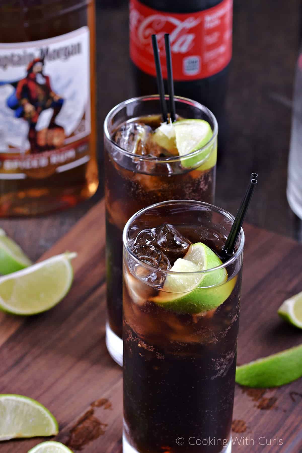 Two tall glasses with rum, cola, ice cubes and a lime wedge surrounded by lime wedges. 