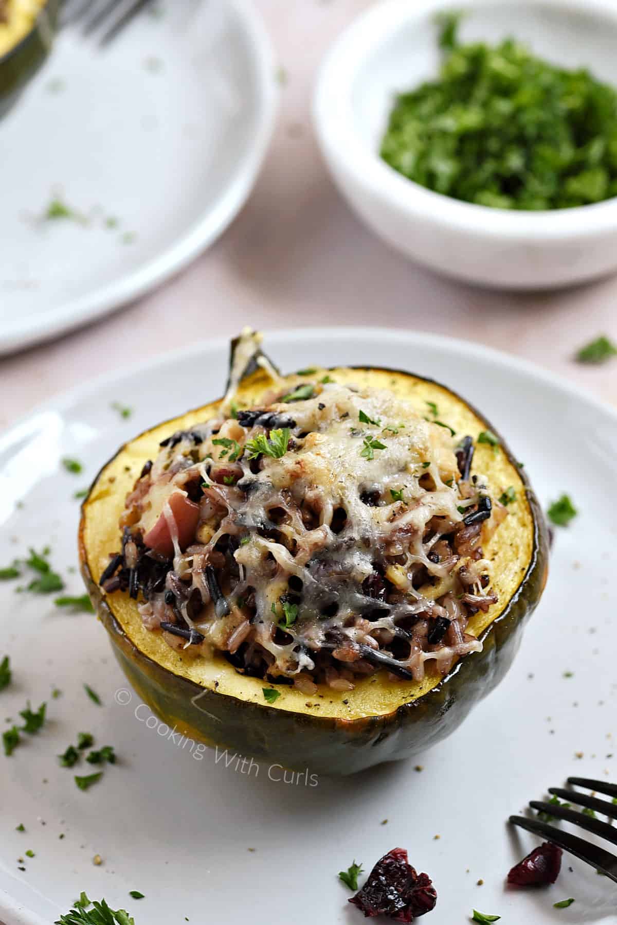 Wild Rice Stuffed Acorn Squash with apples, turkey, and cranberries on a plate with a dish of chopped parsley on the upper right side.