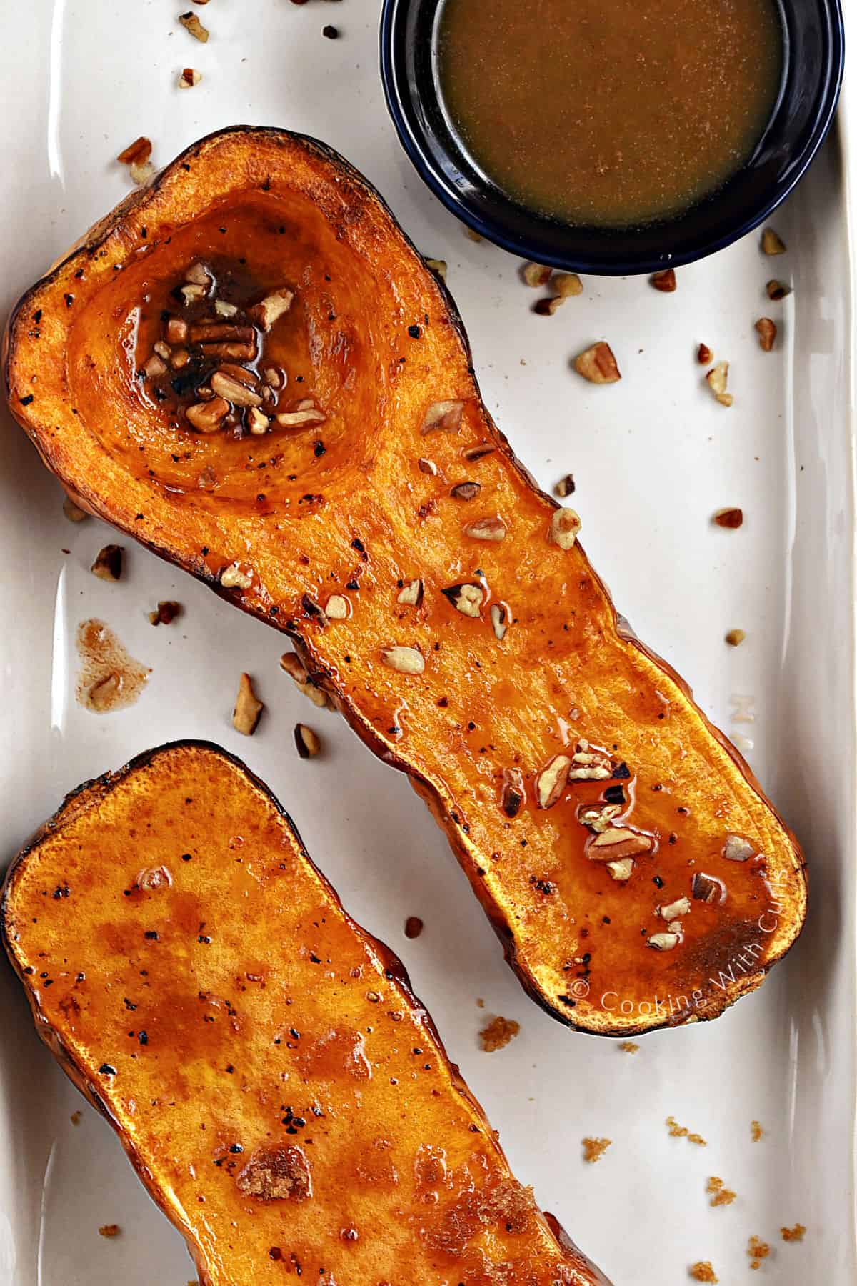 Looking down on two butternut squash halves topped with maple syrup and chopped pecans and butter with brown sugar. 